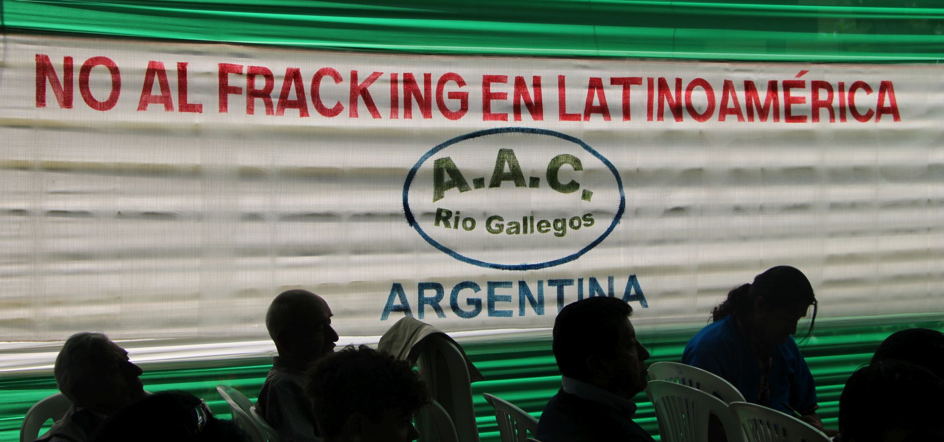 Silhouttes of people sitting in rows in the foreground, in the background a banner with a statement against fracking. 